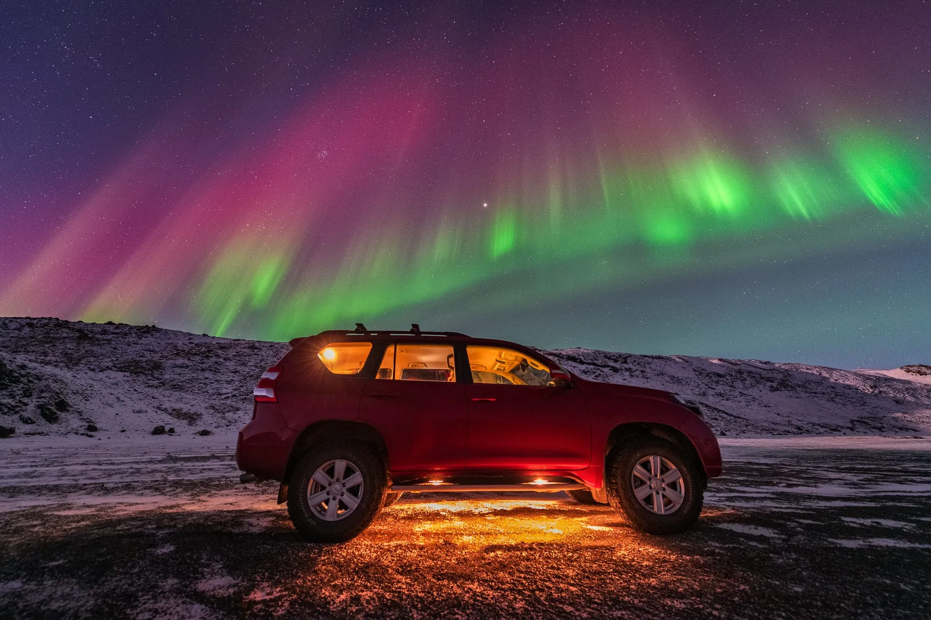รถ SUV สีแดงเปิดไฟจอดบนพื้นที่มีหิมะบางๆ ใต้แสงเหนือสีเขียวและชมพูเต็มท้องฟ้า