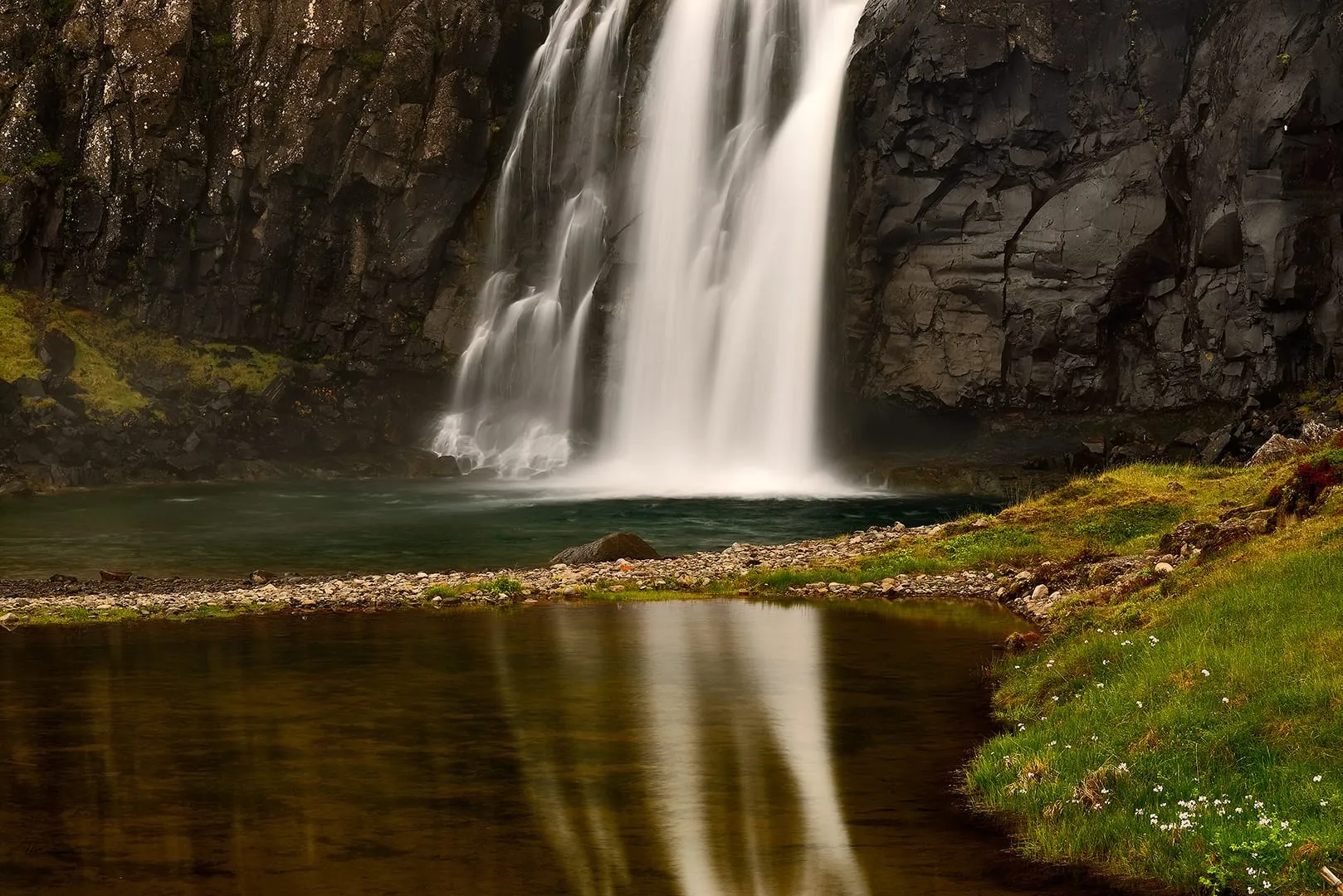 น้ำตกไหลลงแอ่งน้ำใต้หน้าผาหินสีเข้ม เงาน้ำตกสะท้อนในแอ่งน้ำนิ่ง ริมฝั่งมีหญ้าเขียวและดอกไม้สีขาว