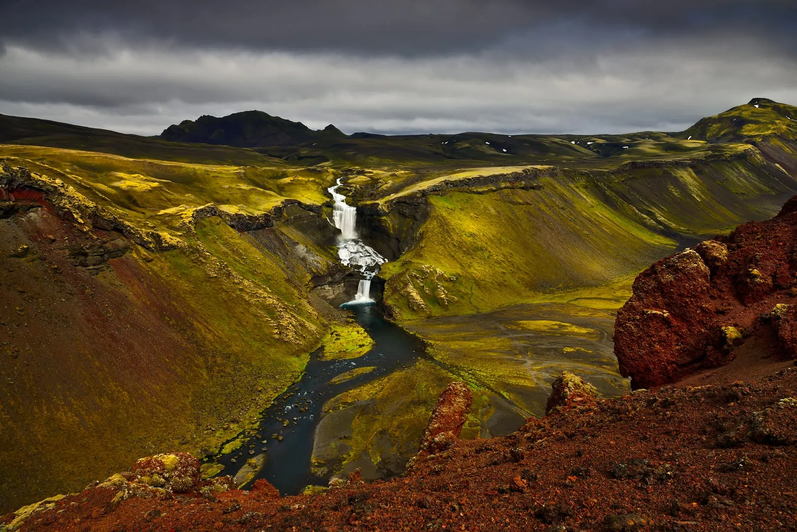 น้ำตก Ófærufoss สองชั้นไหลลงหุบผาสีเขียวมอสใน Eldgjá ด้านหน้าเป็นหินภูเขาไฟสีแดง