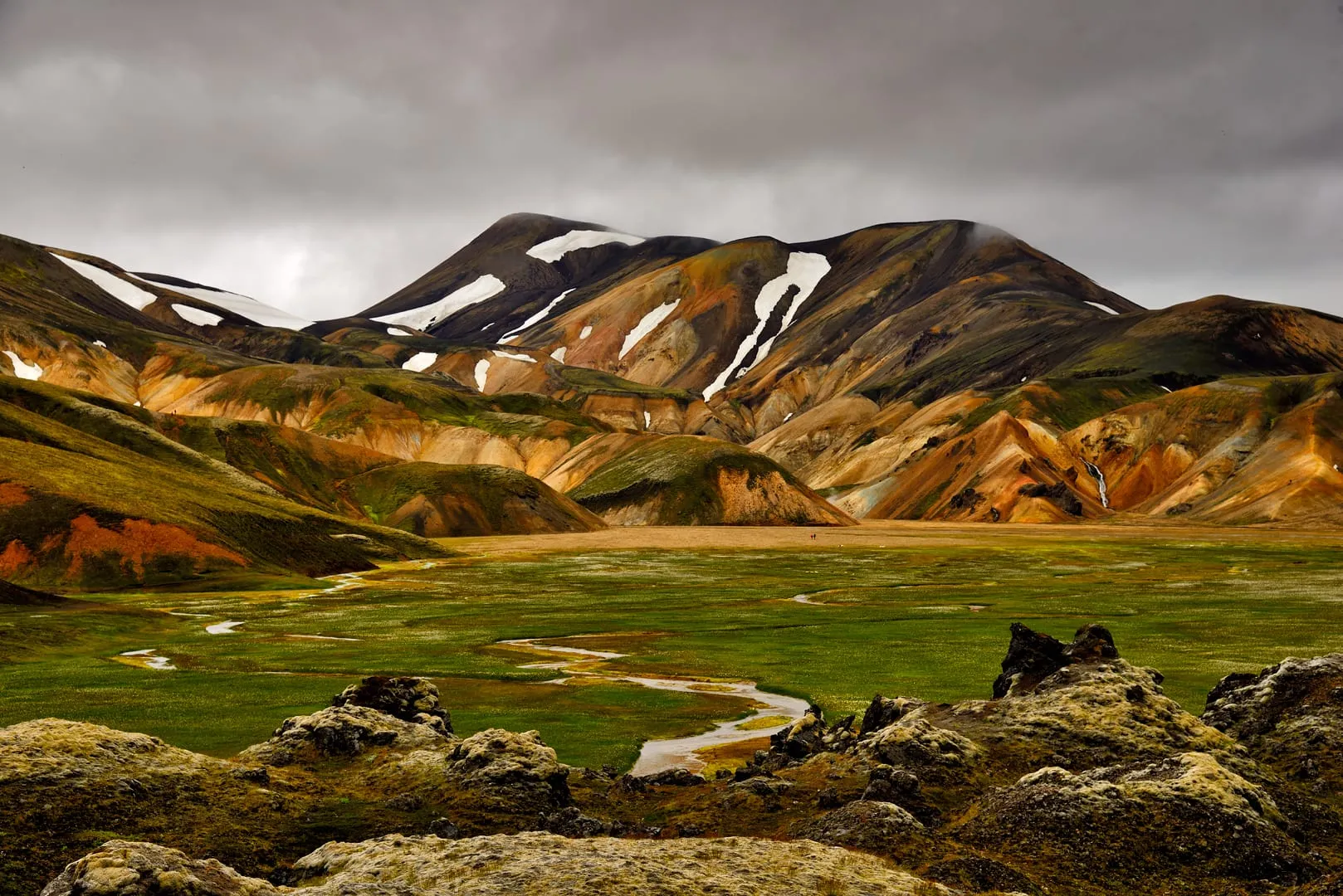 ภูเขาไรโอไลต์หลากสีมีหิมะเป็นแนวที่ Landmannalaugar ทุ่งหญ้าเขียวมีลำธารคดเคี้ยว ด้านหน้าเป็นหินลาวาปกคลุมมอส